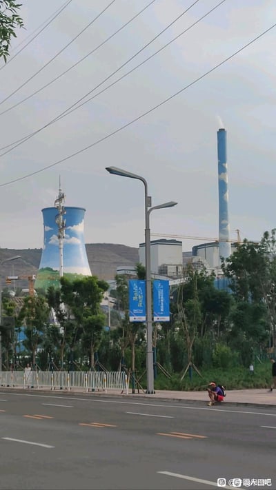 这是一张竖屏实拍的城市街景照片：前景是带有标线的城市道路，路边人行道上有一位行人正坐着休息；中景是行道树、带有蓝色宣传标语的路灯杆；背景是绘有蓝天白云与绿地图案的电厂冷却塔，旁边的蓝色烟囱正排出白色水汽，远处可见山丘与施工塔吊，呈现出工业设施与城市日常结合的平和场景，无谐音梗、双关或暗示内容。