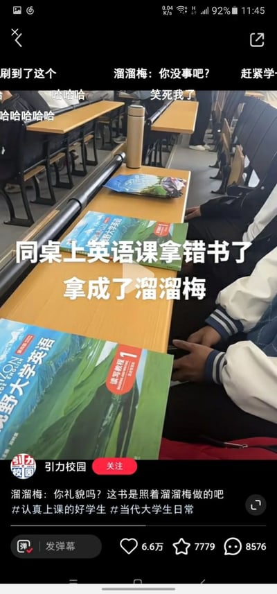这是一张短视频截图，呈现的是大学教室的英语课堂场景。画面中，一名同学在课前准备时，误将溜溜梅的零食包装袋当成了《新视野大学英语读写教程1》课本，因为两者的封面蓝绿配色、画面风格高度相似，闹出了笑话。画面中间配有醒目的白色文字“同桌上英语课拿错书了 拿成了溜溜梅”，顶部有玩梗文字“溜溜梅：你没事吧？”，还有“哈哈哈”“笑死我了”等弹幕烘托搞笑氛围，底部文案则调侃“溜溜梅：你礼貌吗？这书是照着溜溜梅做的吧”，整体充满了轻松搞笑的氛围，精准捕捉了当代大学生校园生活里的趣味小插曲，引发网友对校园日常的共鸣。