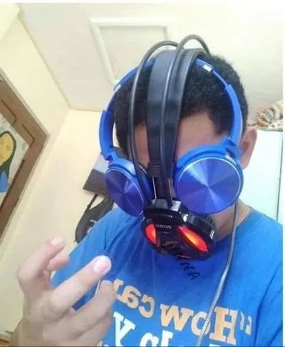 图中男子将三副耳机叠在头上佩戴，一副蓝色头戴式耳机正常架在耳朵上，另外两副耳机（一副黑色头戴式、一副带红色灯光的黑色电竞风格耳机）依次叠在蓝色耳机上方，男子对着镜头比出类似“摇滚”的手势，整个画面十分搞怪，像是在夸张地追求极致的听觉体验，或是单纯为了整活制造趣味效果。