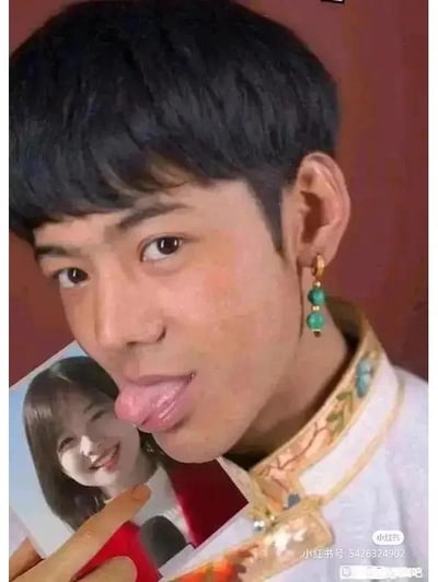 图片中是一位穿着传统服饰的年轻男子，他留着黑色短发，耳朵上戴着绿色玉珠耳环。男子正对着镜头吐舌头，做出搞怪表情，同时用手指着手中拿着的一张女性照片。照片中的女性面带微笑，背景为红色。男子的传统服饰领口有金色刺绣花纹，整体画面呈现出轻松有趣的氛围。