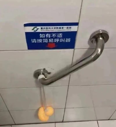 这是重庆医科大学附属第一医院卫生间内的实拍场景：白色瓷砖墙面上贴着蓝底白字的官方提示牌，上方标注医院名称，下方写着“如有不适请按简易呼叫器”，并用红色箭头指向下方的呼叫装置。但令人意外的是，呼叫器的拉绳末端连接的并非常规的呼叫按钮或拉环，而是一双黄色的拖鞋，安装在金属扶手的下方。原本用于紧急求助的医疗设施，却搭配了如此迷惑的拉环设计，形成了强烈的反差感，让人觉得既搞笑又无奈，没有谐音梗或双关含义，单纯是设施设计的不合理带来的趣味效果。
