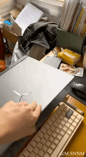 这是一个充满趣味的视觉错位动态图，画面呈现出一个摆放着键盘、笔记本电脑、鼠标、零食杂物等物品的杂乱桌面。一只手看似正准备按下键盘的按键，实际却是按在打开的笔记本电脑的边缘，通过巧妙的拍摄视角，让电脑边缘与键盘按键在视觉上重合，制造出手在按键盘的错觉，带来新奇搞笑的视觉体验。