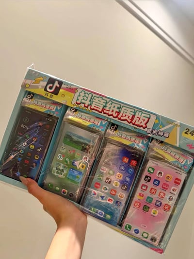 整张图为实拍照片，画面中可见一只手托举着一套全新未拆封的玩具商品，商品外包装印有抖音官方logo，标注有“抖音纸质版 断网神器”的字样，套装内包含4款仿真智能手机造型的纸质玩具，每款玩具表面都印刷了模仿真实手机桌面的各类常用APP图标，涵盖抖音、淘宝、微信、支付宝等常见应用，是主打玩梗属性的整蛊类小商品，调侃用户对短视频APP和手机的依赖，号称断网状态下也能“使用”。