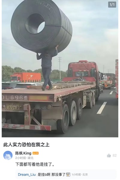 图片中一名男子站在一辆红色卡车的平板上，双手举着一个巨大的黑色钢卷，钢卷直径远超人体高度，看起来异常沉重。男子表情专注，背景是高速公路和其他车辆。图片下方配有文字"此人实力恐怕在我之上"，以及网友评论"下面都写着他是挂了"和"是挂b啊 那没事了"，形成幽默效果。

这张图通过视觉错位和比例反差制造出一种超现实的"大力士"效果，让人产生此人拥有非凡力量的错觉，随后的评论用游戏术语"开挂"来调侃这种不真实的场景，形成搞笑梗。