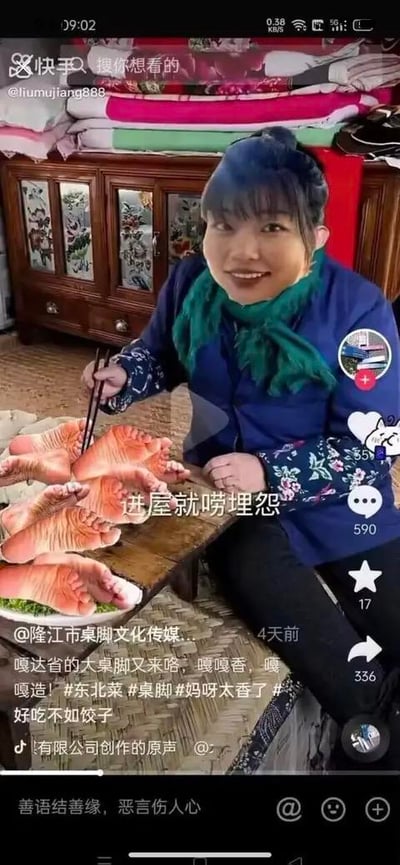 这是一张快手平台的短视频截图，画面呈现了一位身着东北风格服饰的女性，面带笑容坐在桌前，手持筷子做出准备夹取食物的动作。而她面前的“菜肴”是被P图处理成类似猪蹄样式的人的脚掌，制造出荒诞的视觉反差。图中配有文字“进屋就唠埋怨”，下方视频文案用谐音梗“桌脚”替代“猪脚”，搭配东北方言“嘎嘎香，嘎嘎造”，模仿吃东北硬菜的场景进行恶搞整活，文案还附带#东北菜#桌脚#妈呀太香了#好吃不如饺子等话题，整体充满搞怪、荒诞的搞笑氛围，通过视觉和文字的双重反差来制造笑点。