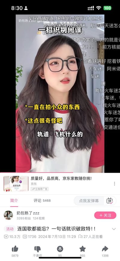 这是一张竖屏的B站移动端视频播放页截图，画面主体是穿红白拼色短袖、戴黑框眼镜的女UP主奶包熟了zzz，画面叠加了黄色字幕“*一直在拍小众的东西 *这点很奇怪吧”和白色字幕“轨道 飞机什么的”，背景还有用户发送的半透明弹幕；页面下方显示该视频标题为“连国歌都能忘？一句话就识破敌特！！”，发布于2024年7月13日，有10.3万播放量、5468条评论，还附带京东家教的推广广告，以及点赞、投币、收藏等B站视频互动按钮。