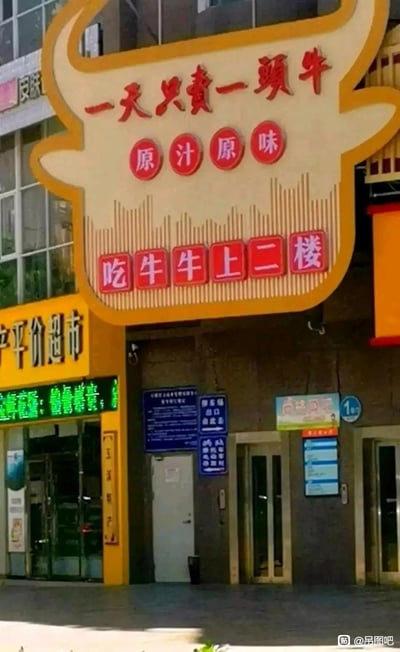 这是一张实拍的街边餐馆招牌照片，招牌设计为牛头造型，上方标注“一天只卖一头牛”“原汁原味”，底部的指引标语“吃牛牛上二楼”因表述方式容易引发奇怪联想，显得格外滑稽。招牌旁还有一家平价超市，整体是日常的城市街边商铺场景。

标语的“吃牛牛”存在歧义，将普通的吃牛肉的指引变得充满搞笑效果。