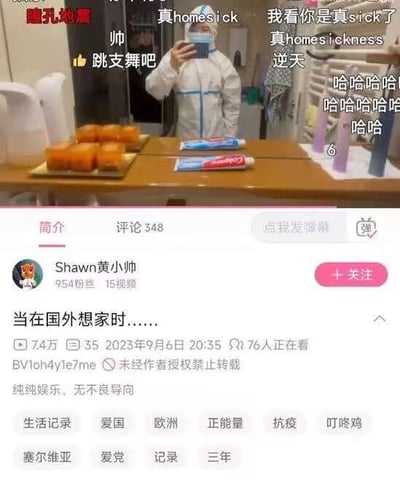 这是B站用户「Shawn黄小帅」的生活记录类视频截图，视频标题为《当在国外想家时……》。画面中博主身着白色抗疫防护服，正对着手机自拍；桌上摆放着橙色房子造型的食物、消毒凝胶和几根试管，呼应了弹幕里的双关梗：英文“homesick”本意为“想家”，被拆分为“home（对应橙色房子）”和“sick（对应防护服、防疫物品，指代隔离生病的状态）”，网友弹幕玩梗“真homesick”“我看你是真sick了”，结合视频主题形成趣味谐音。其他弹幕如“跳支舞吧”“逆天”“哈哈哈哈哈哈哈哈”则是网友的调侃互动。视频简介标注「纯纯娱乐、无不良导向」，标签包含塞尔维亚、抗疫等，推测博主在塞尔维亚处于隔离状态，用这种幽默的方式表达自己想家又身处防疫隔离处境的心情。
