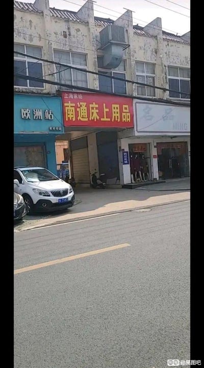 这是一张竖屏拍摄的城镇街景照片，画面展示了一排外墙略显斑驳老旧的临街店铺：左侧是蓝色招牌的“欧洲站”服饰店，中间是红色招牌的“上海家纺 南通床上用品”家纺店，门口立着一块蓝色的“拆迁区域”标识牌，右侧是“名品”服装店，店门口有服装模特。店铺前的空地上停放着两辆汽车，前方是平整的柏油路面，整体呈现出充满生活气息的城镇老街道日常场景，没有双关、谐音梗或暗示内容。