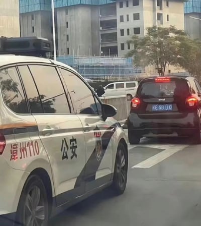 在城市道路的车流中，一辆印有“漳州110”“公安”字样的警车，旁边紧跟一辆黑色smart汽车，其车牌号为“闽E·SB110”，和警车的110标识形成了极具反差感的趣味巧合，仿佛“贴脸开大”，让人忍俊不禁。
该场景的趣味点在于车牌号“SB110”与警车的110标识形成对应，“SB”的谐音效果加上和110的关联，制造出强烈的搞笑反差。