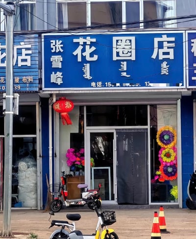 这是一张街边实体店铺的实拍照片，店铺的蓝底白字招牌上竖排标注店主姓名“张雪峰”，横排大字为“花圈店”，同时配有蒙文对照和打码的联系电话。店铺门口悬挂有红色恭喜灯笼，玻璃门窗处摆放着仿真花、花圈等丧葬相关商品，店门前停放着几辆电动自行车，路面还放置了两个橙白相间的交通锥。由于店主姓名和知名网红张雪峰完全相同，网友借这张图玩梗，调侃张雪峰此前停播的事件，配文称他是来这里开花圈店所以没有开直播。