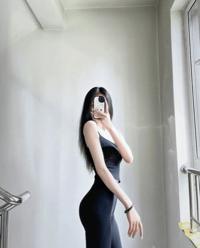 这是一张竖版的原始拍摄静态单图，画面中一位留着黑色长发的女性身处室内靠近窗户的位置，她身着一套黑色紧身运动装，包括吊带款上衣和紧身长裤，身材纤细匀称。她左手拿着手机挡住自己的脸部，右手自然下垂，手腕佩戴着深色手链。背景是浅色的平整墙面、金属栏杆以及一扇窗户，整体环境简约干净，图片风格偏向日常健身穿搭或个人身材状态的展示。