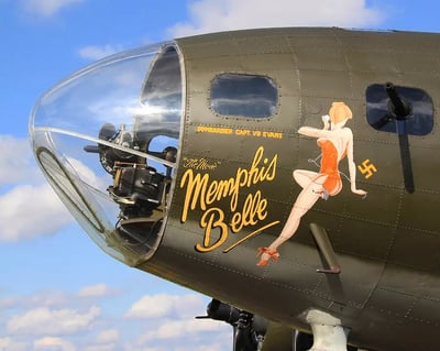图片展示了二战著名轰炸机“孟菲斯美女号”（Memphis Belle）的机头特写。透明的领航员舱罩清晰可见内部设备，机身橄榄绿色金属表面上有两处显著文字：上方为“BOMBARDIER CAPT. VB EVANS”（投弹手VB埃文斯上尉），下方是黄色手写体“The 'Moe' Memphis Belle”。右侧绘有经典的二战机头艺术——一位身着红色紧身衣、脚踩红色高跟鞋的金发女郎，姿态妩媚地侧身倚靠，单手轻扶脸颊。背景是晴朗的蓝天与少量白云，凸显出战机的历史质感与复古美学。