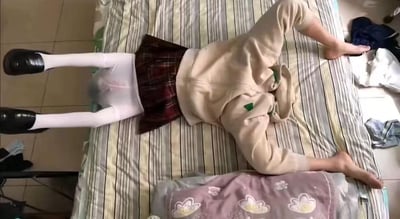 一张俯拍照片，展示一个人以不自然的姿势平躺在床上。床上铺着条纹床单，人物穿着米色连帽衫、红色格子裙、白色长袜和黑色鞋子。人物双腿伸直，双臂向两侧伸展，姿态略显僵硬。床的旁边地板上散落着一些衣物。整个场景看起来像是在卧室中，光线自然。