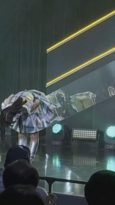 这是一张舞台演出现场的实拍画面，一位穿着银色调演出服、长靴的表演者正弯腰鞠躬，她的裙摆与身后舞台屏幕上的画面恰好重合，形成了仿佛裙摆出现破洞或奇怪图案的视觉错位效果，场景十分滑稽有趣。
无谐音梗、双关或暗示内容，仅为自然形成的视觉错觉。