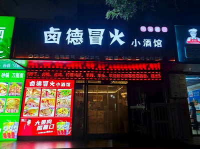 这是一张夜间拍摄的餐馆 exterior 照片，展示了名为“卤德冒火小酒馆”的店铺招牌。主招牌以黑色为底，白色大字突出店名，上方有WiFi、充电等服务标识。招牌下方是红色LED滚动屏，显示“卤味弟弟 绝味鸭货 柠檬鸡爪 下酒菜 烧烤 二楼卡座包厢”等菜品和服务信息。左侧有绿色背景的招牌，包含“砂锅刀削面”等其他品类。店门玻璃上贴有“铜锅”字样，门口LED屏展示多道菜品图片及“大果肉 喝前口”的饮品广告。整体画面灯光明亮，营造出热闹的夜间餐饮氛围。