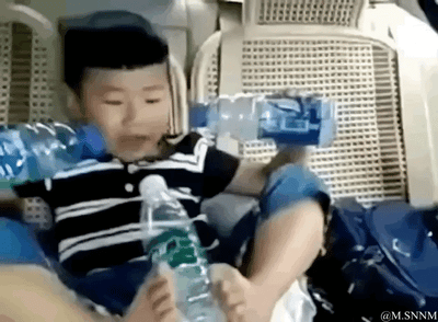 这是一段趣味动态GIF图，画面中的小男孩坐在汽车座椅上，通过巧妙的视觉错位角度，他的两只手和两只脚分别握持着矿泉水瓶，从镜头视角看过去，仿佛小男孩长了四只手同时拿着三个水瓶，制造出充满童趣和搞笑感的视觉效果，让人忍俊不禁。