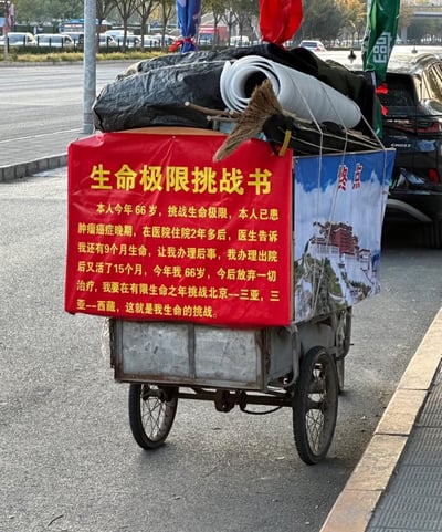 这是一张在城市路边拍摄的实拍照片，主体是一辆改装的三轮车，车上悬挂着红色的《生命极限挑战书》横幅，侧面还有印着布达拉宫图案的“终点”宣传布，同时装载着行李、扫帚等生活物资。横幅上的文字讲述了一位66岁的癌症晚期老人的经历：他曾被医生告知只剩9个月生命，办理出院后却又存活了15个月，如今他放弃一切治疗，计划用剩余的生命完成从北京到三亚、再从三亚到西藏的长途旅行挑战，以此对抗命运，诠释生命的韧性。