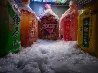 这张图拍摄于冰箱冷冻层内部，画面中整齐排列着五瓶不同口味的康师傅大瓶装饮料，从左至右依次是低糖款绿茶、茉莉蜜茶、热带风味冰红茶、冰糖红西柚、冰糖雪梨。瓶身表面和周围的空间都覆盖着厚厚的冰霜，部分饮料瓶身因内部液体结冰膨胀，冰霜在瓶口和瓶身堆积，营造出一种饮料被“冰封”的趣味场景，视觉上充满冰爽感，仿佛能传递出冷冻环境的低温氛围。