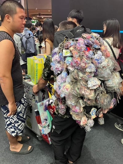 这张照片拍摄于某动漫展现场，画面中心是一位背着巨型背包的男性，背包表面密密麻麻挂满了数百个不同款式的动漫角色钥匙扣和徽章，形成极具视觉冲击力的“周边铠甲”。周围可见众多戴着口罩的参展人群，有人手持印有“BOOCH THE HEROES”字样的黄色购物袋，整体场景热闹拥挤，充满二次元文化氛围。