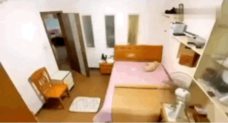 这是一段动态GIF，展示了一个狭小的出租屋内部场景。画面中利用视觉错位效果，让房间里的纸箱在特定视角下看起来像是床铺，当镜头移动后，错觉被打破，才看清是堆叠的纸箱。房间内还摆放着木椅、小床头柜、简易床架，整体布置简单朴素，这个视觉错位为日常的出租屋场景增添了趣味感，让人产生一瞬间的错觉。