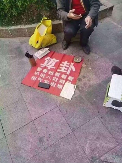 一位 elderly 男性蹲坐在人行道边摆摊算命，面前铺着一块红色布幔，上面用白色大字写着“算卦”及多项服务内容：看手相、看运气、批八字、看财运、看姻缘。布幔周围摆放着几本旧书、一个插着笔的笔筒和一个黄色布袋，整体场景呈现出传统街头算命的典型 setup，人物神态平静，似乎在等待顾客。