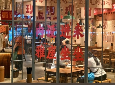 这张图片展示了一家中式复古风格餐厅的外观，透过玻璃门可以看到内部座位和顾客。玻璃门上贴着大红色的中文字：“内设雅座 进来坐坐”，氛围热闹而温馨。店内灯光柔和，墙上悬挂着复古广告海报和菜单，桌椅为木制。图中可见顾客正在用餐或休息，左边有穿工作服的员工。

未见明显双关、谐音梗或暗示。画面传达了邀请和轻松氛围。