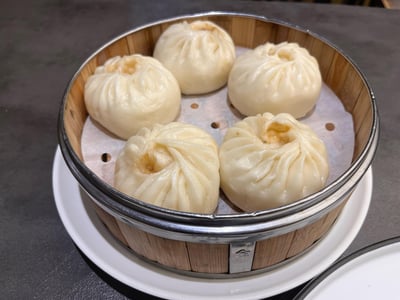 一张实拍的美食照片，竹制不锈钢包边的蒸笼里摆放着五个白白胖胖的蒸包，包子顶部褶皱清晰，收口处带着些许油润的色泽，蒸笼底部垫着带孔蒸纸，放在白色瓷盘上，背景是深灰色的桌面，看起来松软可口，是典型的中式早餐点心。