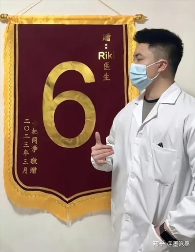 画面中，一位佩戴蓝色口罩、身着白色医生服的男士站在墙边，左手插兜，右手比出模仿数字6的手势，侧头看向身旁墙上悬挂的锦旗。锦旗主体为酒红色，边缘是金色装饰，中间印有醒目的金色大数字“6”，上方标注“赠：Riki医生”，左下角则是“小把同学 敬赠 二〇二三年三月”的字样。这里的数字6是网络热词“666”的简化用法，用来表达对医生医术或服务的夸赞，意为“厉害、牛”，结合医生摆出的同款手势，形成了趣味玩梗的场景，充满了轻松搞笑的氛围。