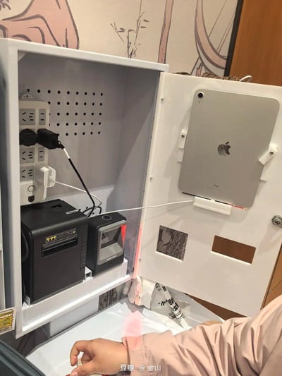 这是一张店铺收银场景的实拍图，展示了一个整合式收银设备收纳柜：柜门内侧通过定制支架固定着一台银色iPad，正通过白色充电线连接柜内的多口插座进行充电；柜内左侧安装了带多个插孔的电源插座，连接着黑色小票打印机和带红光提示的扫码设备的供电线，两台设备被放置在柜内的分层架上；柜内下方还能看到一些包装用品，画面下方露出了一只手的部分。这个收纳柜将收银相关的电子设备集中收纳，既节省空间又让设备布局更整洁有序。