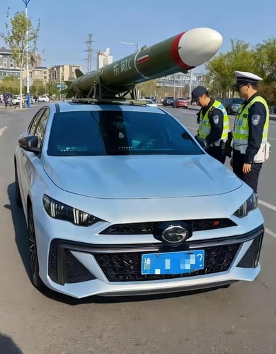 在城市的开阔道路上，一辆浅蓝色的广汽传祺影豹R轿车被车主违规加装了一个标注“东风1C”的大型仿真导弹模型，放置在车顶位置。两名身着反光执勤服的交警正在对该车辆进行检查，一名交警低头查看相关材料，另一名在旁观察。这种普通家用车搭配军事装备模型的改装充满荒诞搞笑的反差感，因属于违规改装车辆，所以被交警依法拦截检查。