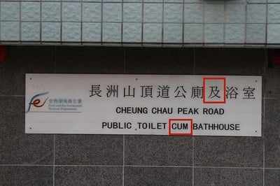 图片展示了香港长洲山顶道一处公共设施指示牌，牌上中文为“长洲山顶道公厕及浴室”，英文翻译为“CHEUNG CHAU PEAK ROAD PUBLIC TOILET CUM BATHHOUSE”。其中“及”被翻译为“CUM”，而“cum”在现代英语中常带有粗俗含义，引发翻译不当的争议。指示牌左侧有“康乐及文化事务署”的中英文标识，背景为灰色瓷砖墙面。
