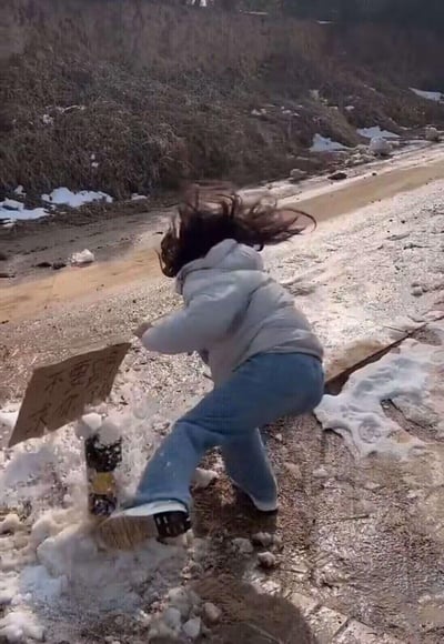 图片中一名身穿灰色连帽卫衣和蓝色牛仔裤的女生正在乡村土路上愤怒地踩踏一个立在雪堆中的警示牌。她的头发因动作而飞扬，右腿高高抬起，正用力踩向写有「不要踩我」字样的木板牌。周围环境显示这是冬季，地面有未融化的积雪和结冰，背景是长满枯草的山坡。画面充满戏剧性，形成了「越是警告不要踩反而越要踩」的讽刺效果。