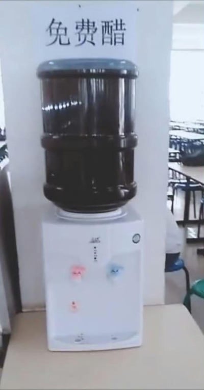 一张在食堂或餐厅拍摄的照片，画面中央是一台白色的饮水机，上面放置着一个黑色的大水桶。饮水机上方的墙壁上贴着一张写有“免费醋”三个黑色大字的纸张，将原本应该装饮用水的饮水机改造成了提供免费醋的装置，形成一种有趣的反差感。