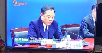 淄博新闻综合频道播出的会议画面，市自然资源和规划局党组书记、局长汪德法正在发言。画面中汪德法身着蓝色西装，坐在会议桌后，面前有麦克风和名牌。背景为蓝色，显示'新闻综合'和'淄博新闻'标识。