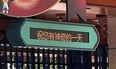 这是一张实拍场景图，画面主体为一个带有青绿色装饰边框的电子显示屏，屏幕以像素风格的字体呈现出温馨祝福语“祝您有神奇的一天”。显示屏被悬挂在空间上方，其下方是一排带有玻璃门的深色格子柜，柜内摆放着色彩各异、带有图案的物品，看起来像是主题文创周边。显示屏上方还露出了一部分带有卡通形象的圆形装饰，整体场景充满童趣与温馨感，推测是主题商店或游乐园区内的场景，传递出愉悦、梦幻的氛围。