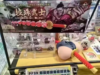 图片展示了一个商品陈列场景，核心是一款名为“拉珠武士”的玩具包装。包装上印有肌肉发达的武士风格角色插画，角色表情狰狞，手持带有红色珠子的武器。包装上有中日双语文字，主要内容包括“拉珠武士DX天诛丸×台湾知名大便绘师【麒麟】”“拉珠数8颗”“全长52.3cm”等信息，还印有“ここには武士がいない、情けない!!”（这里没有武士，真可悲!!）的日文标语。包装下方的展示台上摆放着该玩具的实物，可见红色手柄和蓝色布料装饰的部件。整体风格猎奇搞笑，属于 niche 玩具产品展示。