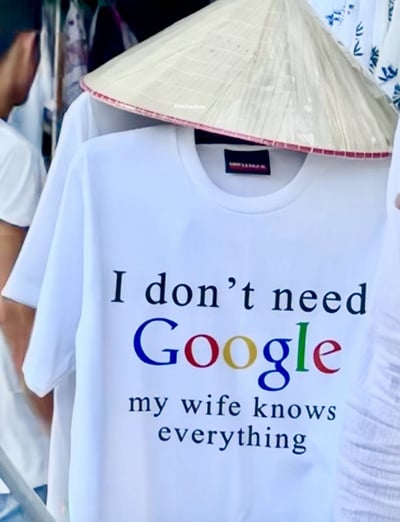 这是一张拍摄于市集或街头摊位的T恤图片，T恤正面大字印着："I don't need Google, my wife knows everything"。其中"Google"用谷歌标志配色，非常有辨识度。整体表达幽默戏谑，意思是“有了老婆，就不需要查Google，因为老婆什么都知道”，带点自嘲，也包含夸赞和温情意味。

有轻松夫妻关系的梗味，同时Google的logo色配也让文字很抓眼。斗笠帽的出现，让画面带有越南乃至东南亚集市的生活气息。