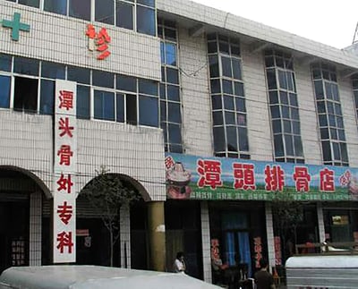 这是一张城镇街景实拍图，画面中的临街建筑被分为两家店铺：左侧是带有绿色十字标识的「潭头骨外专科」诊所，门脸为拱形设计，竖排招牌醒目；右侧紧邻着「潭头排骨店」餐馆，招牌上印有菜品图案，主打排骨类餐食。两家店铺名字高度相似，一个是骨科医疗诊所，一个是主打排骨的餐馆，这种相邻的搭配形成了有趣的巧合，容易引发联想。画面下方可见部分车辆和少量行人，整体呈现出日常的城镇商业街氛围。