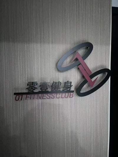 图片展示了一块木质纹理墙面上的健身俱乐部招牌。招牌主体为“零壹健身”四个黑色立体汉字，下方配有红色英文“01 FITNESS CLUB”。右侧是品牌标志，由两个黑色椭圆形环交叉组成，环内有一个红色的箭头状图形，整体设计简洁现代。