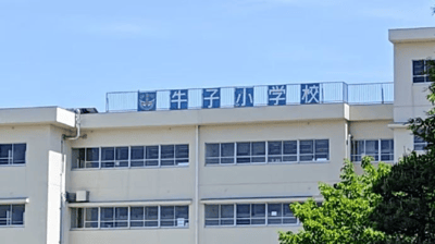 图片展示了一栋两层高的浅色教学楼，建筑外墙上有整齐排列的玻璃窗，顶部天台处悬挂着蓝色底白色文字的校牌，内容为“牛子小学校”。背景是晴朗的蓝天，右下角可见部分绿色树木，整体画面清晰展现了学校建筑的日常外观。