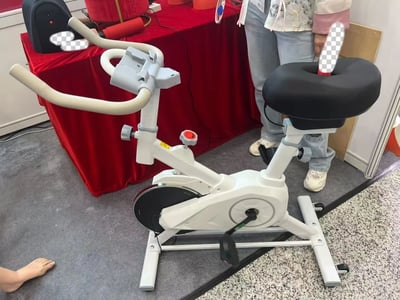 图片展示了一台在展览中展出的健身自行车，其座椅设计较为独特，呈黑色宽大圆形。自行车主体为白色，配有灰色扶手。背景中有一张铺着红色桌布的展台，上面摆放着黑色背包、红色圆柱形物品等。一名穿着浅蓝色牛仔裤和粉白相间上衣的人站在自行车旁，部分手部和物品被带有透明格子的模糊处理。整体场景像是在展会或产品展示现场，地面为灰色地毯，右侧可见部分大理石纹理地面。