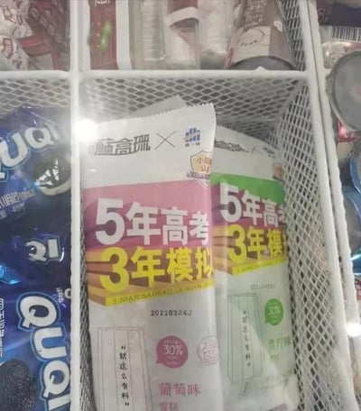 这是一张在超市/便利店冰柜中的实拍图，画面主体是两款「五年高考三年模拟」联名雪糕，分别为葡萄味和青柠味。雪糕的包装高度还原了知名高考教辅《五年高考三年模拟》的封面设计风格，熟悉的「5年高考 3年模拟」字样醒目，搭配「就这么有料」的标语，还标注了30%的果味含量。冰柜中还能看到旁边的奥利奥风格雪糕，整体场景充满了反差感——原本是学生时代备考的“硬核”教辅，摇身一变成了可以食用的趣味雪糕，用IP情怀制造出新奇搞笑的效果，很容易引发学生群体的共鸣。