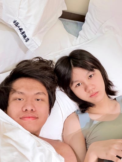 这是一张真人实拍的竖版静态照片，采用俯拍视角，画面中一男一女两名年轻人并排躺在酒店的床上，两人的头发都略显凌乱，共同面对镜头表情自然放松，床上的枕套上印有“泊乐·云景酒店”的字样，整体场景是二人在酒店休息时拍摄的自拍，配合标题调侃了“早上睡觉晚上起床”的颠倒作息。