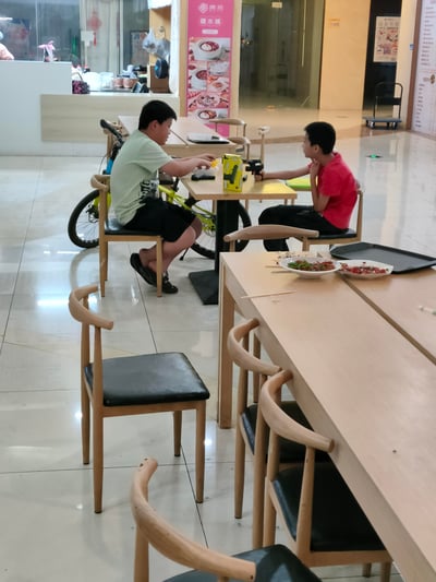 这是一张竖屏的原始生活拍摄图，场景位于一家室内餐厅。画面中，两个小男孩坐在餐桌旁：左侧穿浅绿上衣黑短裤的男孩正专注地摆弄桌上的物品，旁边靠着一辆黄绿配色的儿童自行车；右侧穿红色上衣黑裤子的男孩手持玩具枪，一只手放在脖子处，看向对面的男孩。前景的餐桌上摆放着吃剩的餐盘，背景可见印有“暖燕糖水铺”字样和糖水菜品图的宣传立牌，整体呈现出日常休闲的儿童玩乐场景，没有双关、谐音梗或暗示内容。