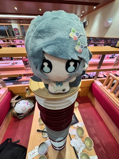 图片展示了寿司郎（スシロー）餐厅内的场景，一个蓝色头发、带有彩色花朵发饰的毛绒玩具被放置在一摞高高堆叠的红白黄三色碗顶上。毛绒玩具有着大大的黑色眼睛和呆萌的表情，脖子系着蓝色蝴蝶结。背景可见餐厅的传送带、餐桌以及其他用餐的顾客，桌面上散落着一些空碗和一个带有卡通贴纸的头盔。整体氛围轻松有趣，展现了在寿司店用餐后堆积的碗与可爱毛绒玩具的趣味组合。