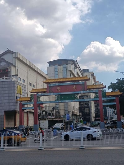 这是一张户外拍摄的日常城市街景图，画面核心是一座红柱黄顶、带有中式传统建筑风格的“唐人街”牌坊，牌坊上的金色字体醒目。牌坊后方是几栋高低错落的现代楼宇，左侧建筑外立面上有麦当劳的经典黄色M标识，下方是24小时营业的门店入口。画面下方的道路区域，有白色护栏分隔，护栏内可见白色轿车、黄色出租车，还有一位骑行电动车的路人。天空中点缀着大朵蓬松的白云，整体氛围平和，是普通的城市日常场景，没有双关、谐音梗或特殊暗示内容。