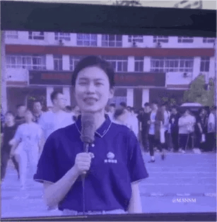 这是一张直播画面的屏幕截图，画面中一位身穿深蓝色带媒体标识上衣的女记者手持话筒，面带笑容，正在进行现场报道。背景是一所学校的户外场地，大量学生模样的人群聚集在此，后方是学校的教学楼，整体场景处于校园内的活动现场，氛围热闹鲜活。