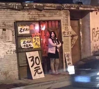 这是一张充满趣味的视觉错位照片，拍摄于夜晚的街边简陋小店门口。一位女士抱臂站在店门口，门口立着一块写有“30元”的大木牌，木牌的位置恰好和女士的下半身重合，从特定角度看仿佛女士的下半身就是这块“30元”的牌子，营造出荒诞搞笑的效果。店铺门口还贴着多张写有“老太太 三十元”“30元”的纸张，墙面显得陈旧，充满市井生活的烟火气。
