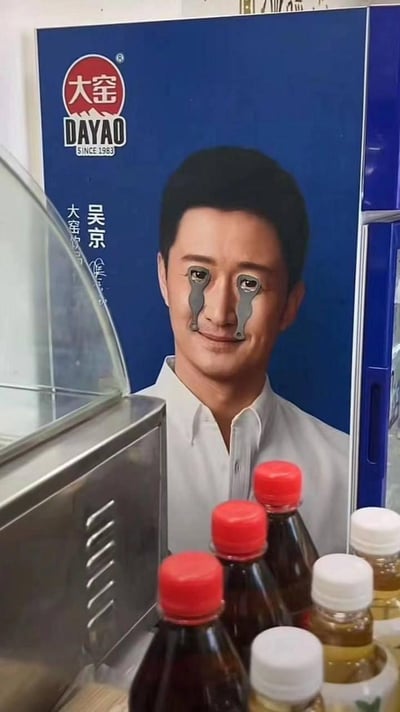 这是一张线下实拍的趣味错位图：大窑饮料的吴京广告立牌前，两个大窑饮料的金属开瓶器刚好卡在立牌上吴京的眼睛位置，形成类似机械义眼的滑稽视觉效果。立牌下方摆放着多瓶大窑饮料和其他饮品，场景看起来是在小卖部或餐饮店内。
没有双关、谐音梗，完全是巧合的视觉错位带来的搞笑效果。